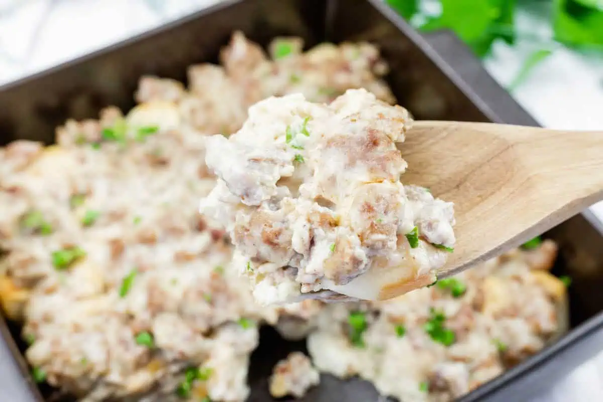 A wooden spoon holds a serving of biscuits and gravy with sausage, over a baking dish filled with the same dish, garnished with chopped herbs.