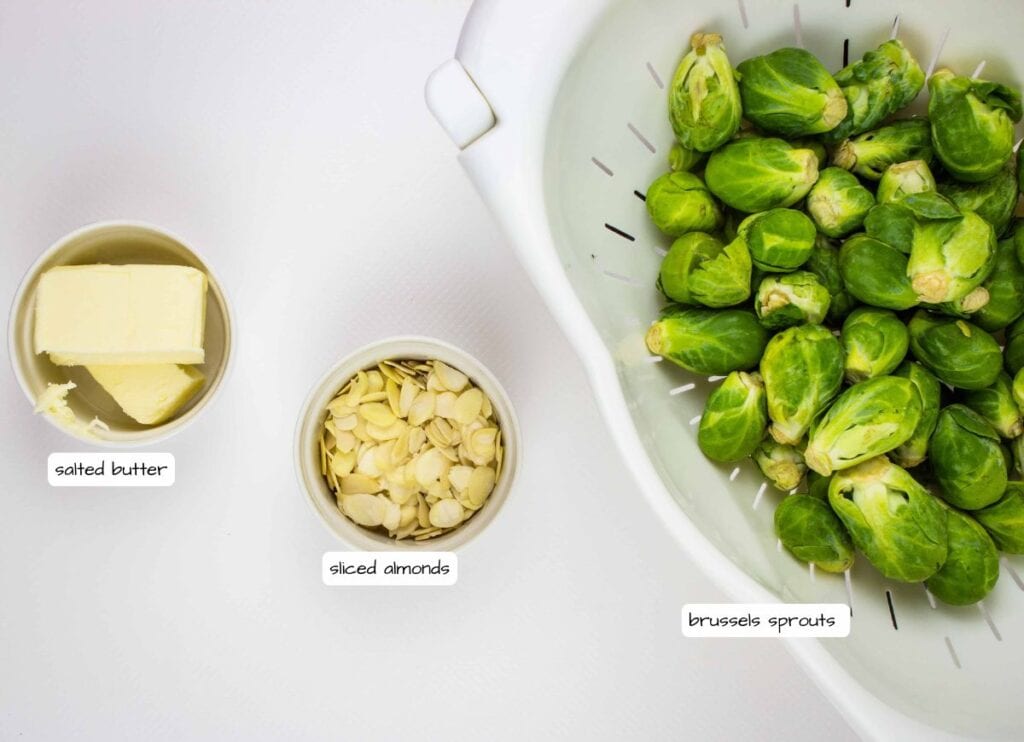 A bowl of salted butter, a bowl of sliced almonds, and a colander filled with brussels sprouts are arranged on a white surface.