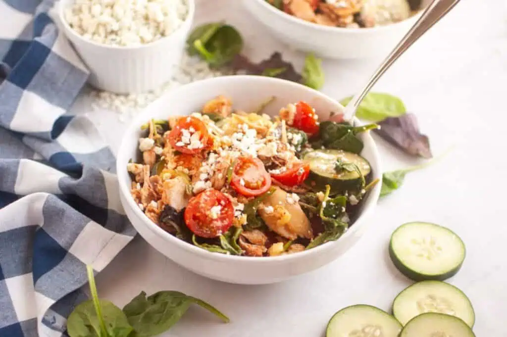 A bowl of salad with cherry tomatoes, cucumbers, spinach, chicken, and crumbled cheese, next to a blue checked napkin and sliced cucumber on a white surface.