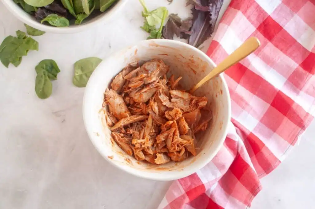 A bowl of shredded, seasoned jackfruit with a gold spoon, next to a bowl of mixed greens and a red checkered cloth on a white surface.