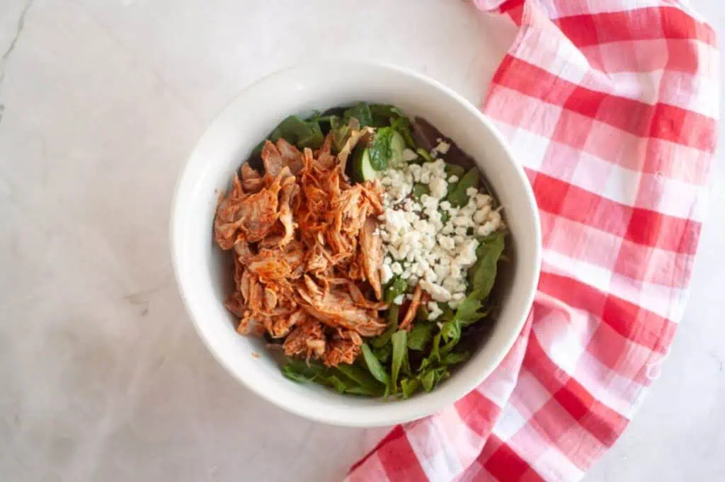 A white bowl containing shredded chicken, mixed greens, and crumbled cheese on a marble surface with a red and white checkered cloth beside it.