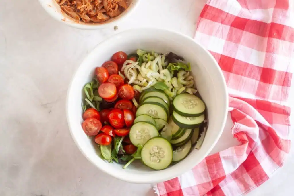 A white bowl filled with sliced cucumbers, cherry tomatoes, celery, and mixed greens sits next to a red and white checkered cloth on a light surface.