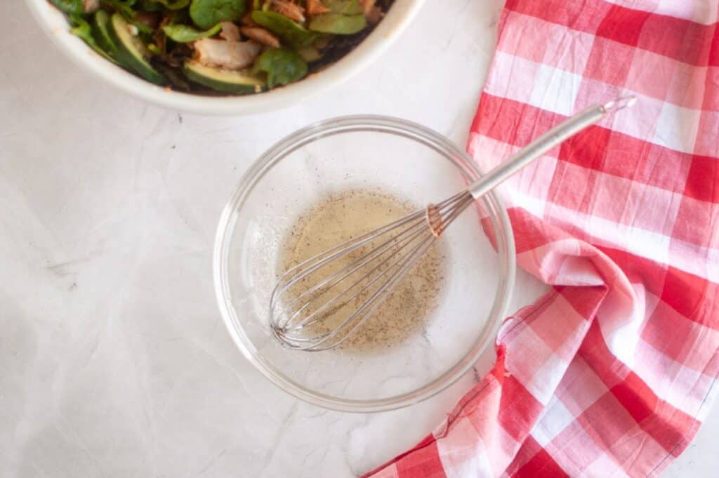 A glass bowl with whisked vinaigrette and a metal whisk beside a red and white checkered cloth; a salad bowl is partially visible in the corner.