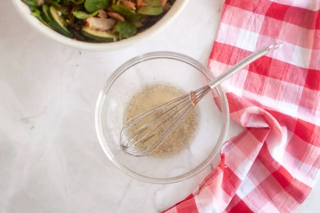 A glass bowl with whisked vinaigrette and a metal whisk beside a red and white checkered cloth; a salad bowl is partially visible in the corner.