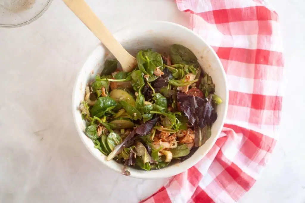 A white bowl filled with mixed greens, shredded meat, and sliced vegetables, with a wooden spoon, sits on a marble surface next to a red and white checkered cloth.