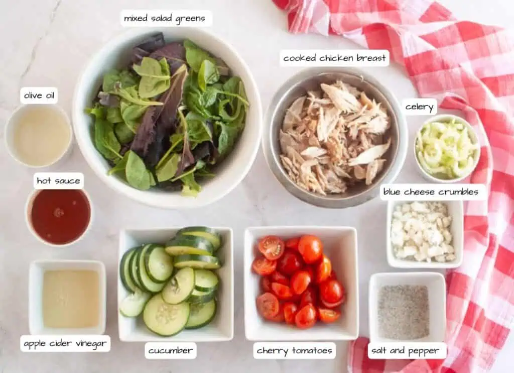 Bowls and dishes with mixed salad greens, shredded chicken breast, celery, blue cheese crumbles, cherry tomatoes, cucumber, olive oil, hot sauce, apple cider vinegar, and salt and pepper.