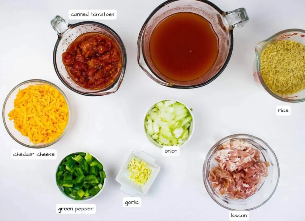 A top-down view of measured ingredients in bowls, including canned tomatoes, chicken broth, rice, cheddar cheese, onion, green pepper, garlic, and bacon, labeled accordingly.
