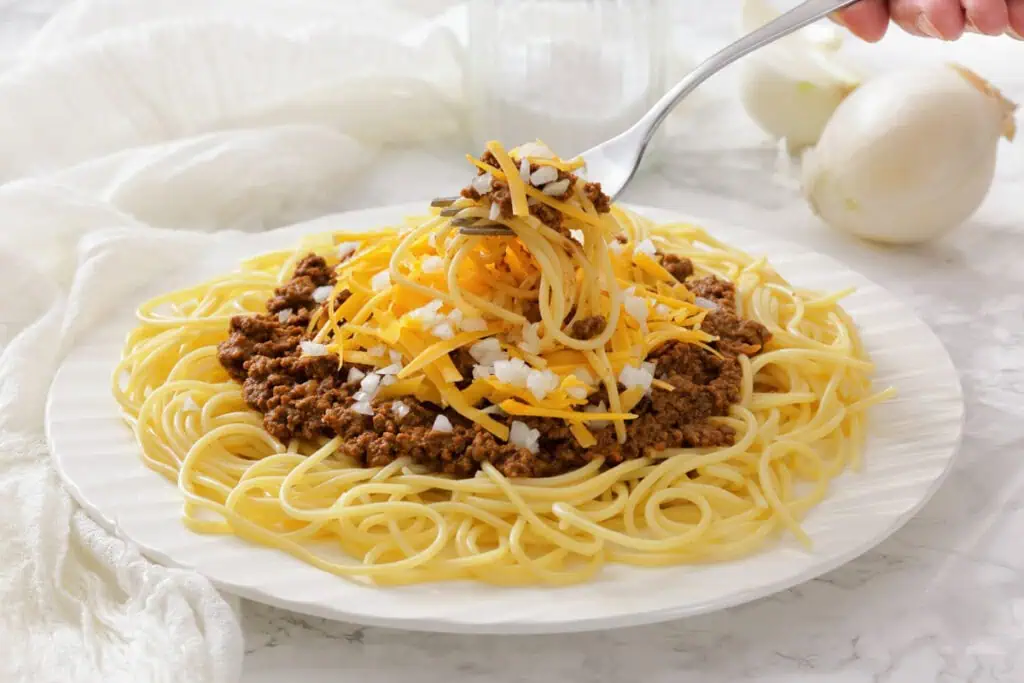 A plate of spaghetti topped with chili, shredded cheddar cheese, and chopped onions, with a fork lifting a portion.