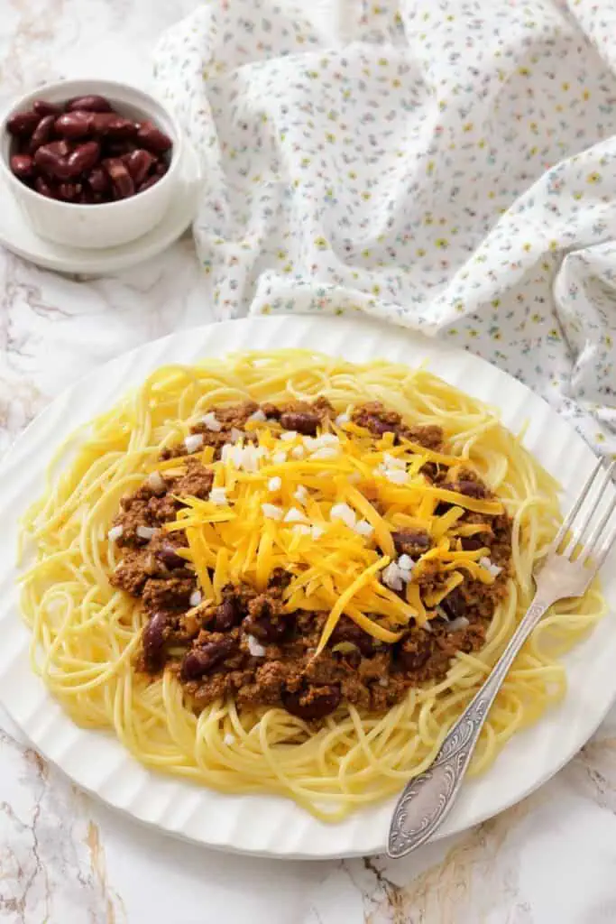 A plate of spaghetti topped with chili, shredded cheddar cheese, and diced onions, with a fork on the side and a bowl of kidney beans in the background.