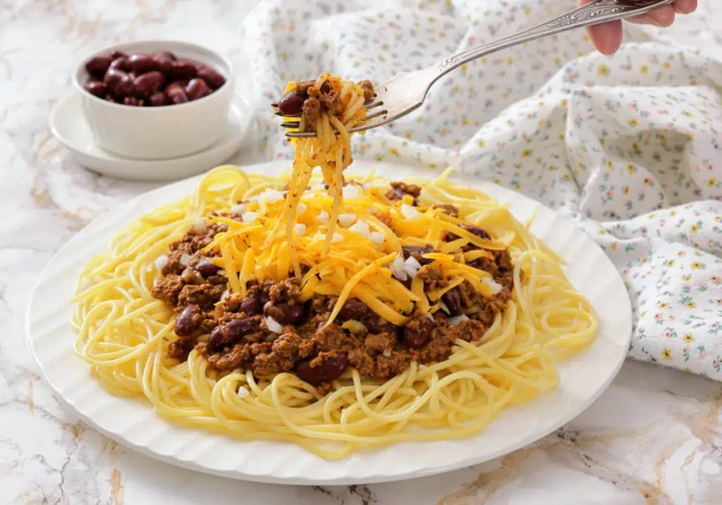 A plate of spaghetti topped with chili, shredded cheddar cheese, and chopped onions, with a fork lifting a bite. A small bowl of kidney beans is in the background.
