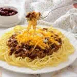 A plate of spaghetti topped with Cincinnati chili, shredded cheddar cheese, and chopped onions, with a fork lifting a bite.