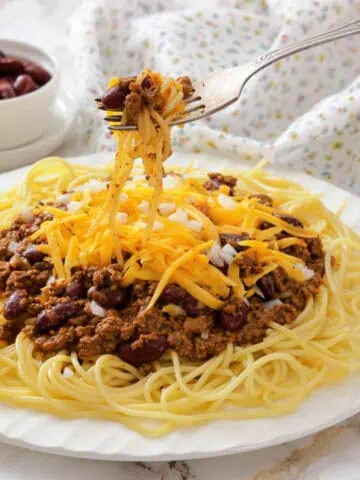 A plate of spaghetti topped with Cincinnati chili, shredded cheddar cheese, and chopped onions, with a fork lifting a bite.