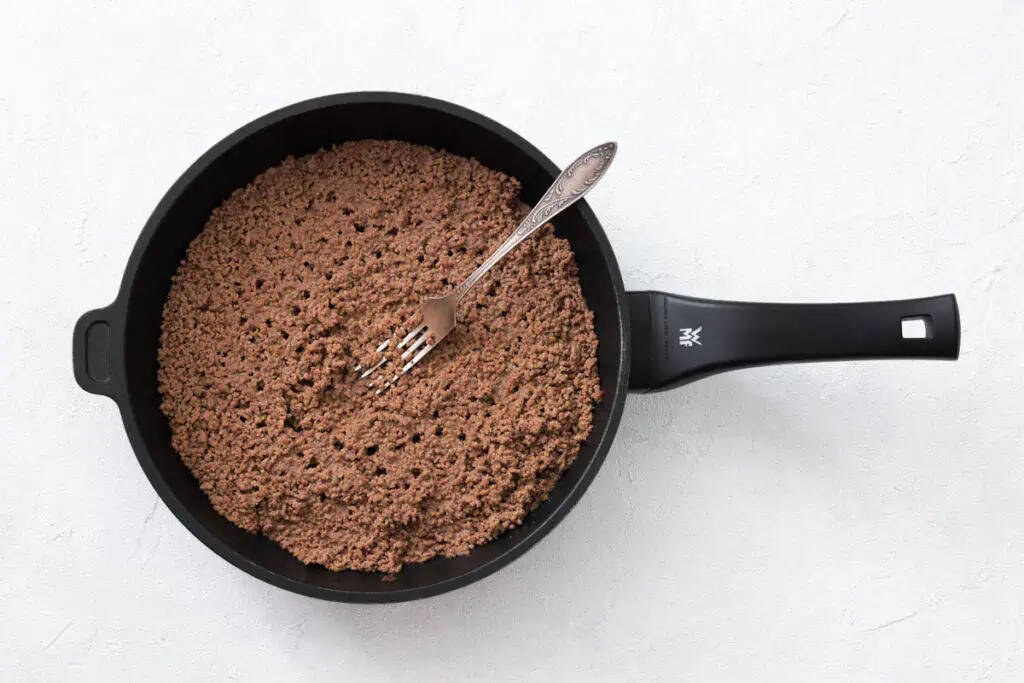 A pan filled with cooked ground beef sits on a light surface; a metal fork rests inside the pan.
