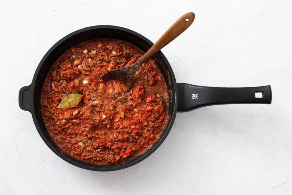 A black saucepan filled with red meat sauce, diced vegetables, and a bay leaf, with a wooden spoon resting inside, on a white surface.
