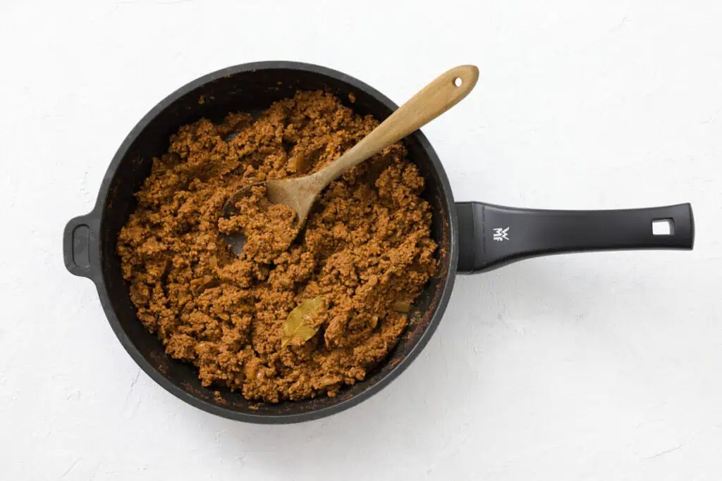 Cooked ground meat mixture with a bay leaf in a black frying pan, stirred with a wooden spoon, on a white background.