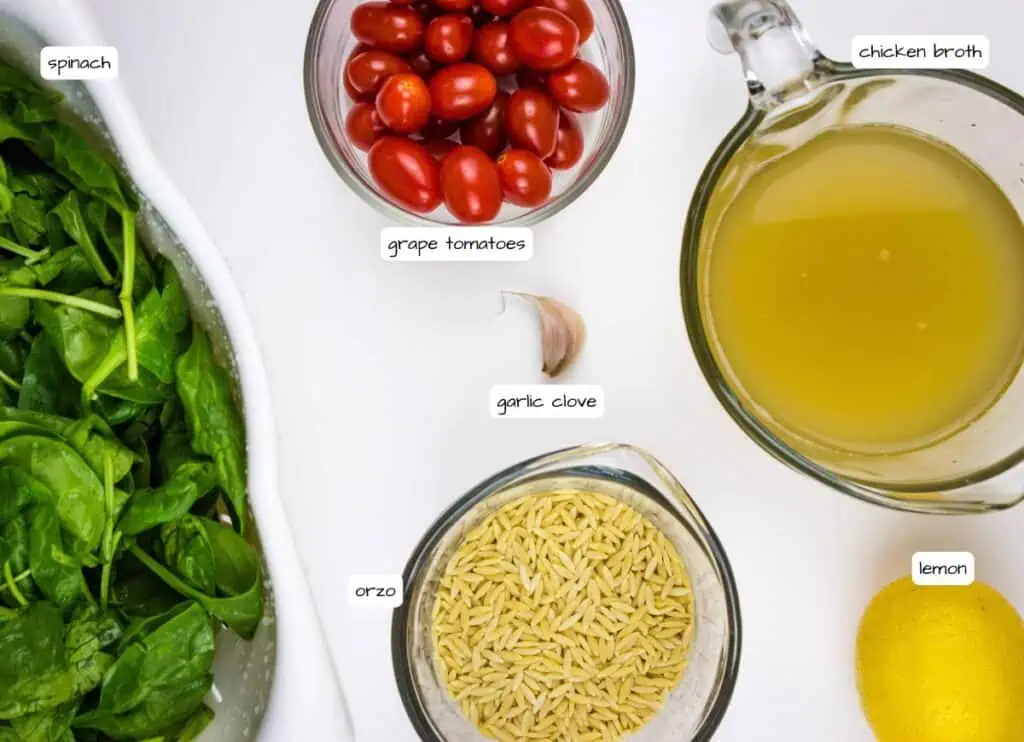 An overhead view of spinach, grape tomatoes, a garlic clove, chicken broth, orzo, and a lemon arranged on a white surface, each labeled with text.