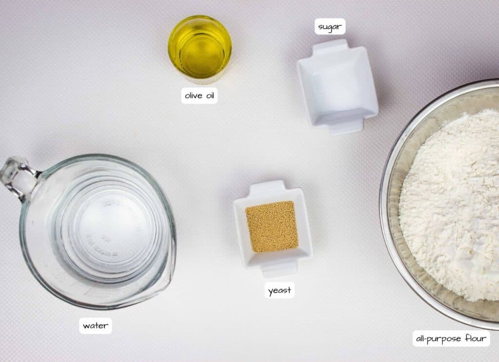 Top-down view of measured ingredients for baking: a bowl of flour, a glass of water, a small bowl of olive oil, and two dishes containing sugar and dry yeast, all labeled.