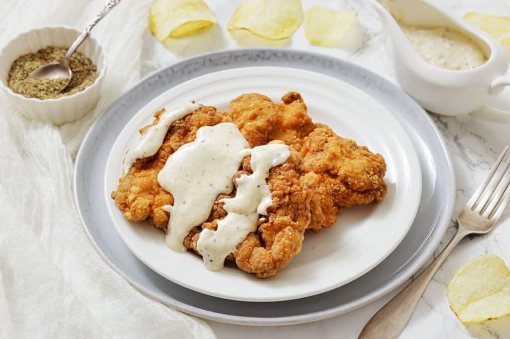 A plate of fried chicken steak topped with white gravy, served with a fork and pepper in a small bowl on the side.