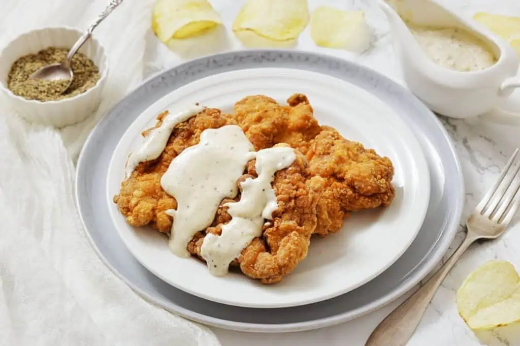 A plate of fried chicken steak topped with white gravy, served with a fork and pepper in a small bowl on the side.