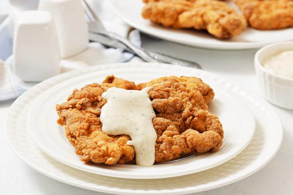 A plate of chicken fried steak topped with white gravy, with another plate and utensils in the background.