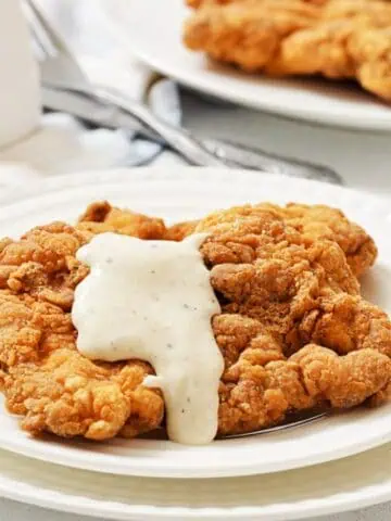 A plate of chicken fried steak topped with white gravy, with another plate and utensils in the background.
