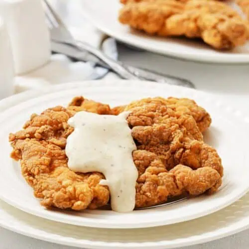 A plate of chicken fried steak topped with white gravy, with another plate and utensils in the background.