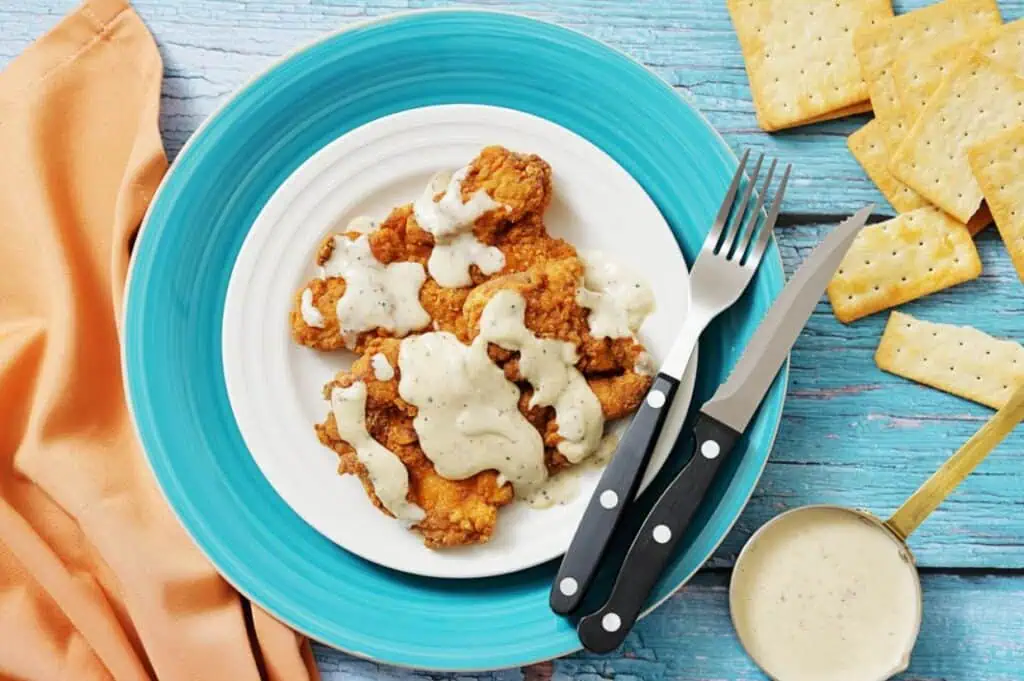 A plate of fried chicken strips topped with white gravy, next to a knife, fork, crackers, a small bowl of gravy, and an orange napkin on a blue wooden surface.