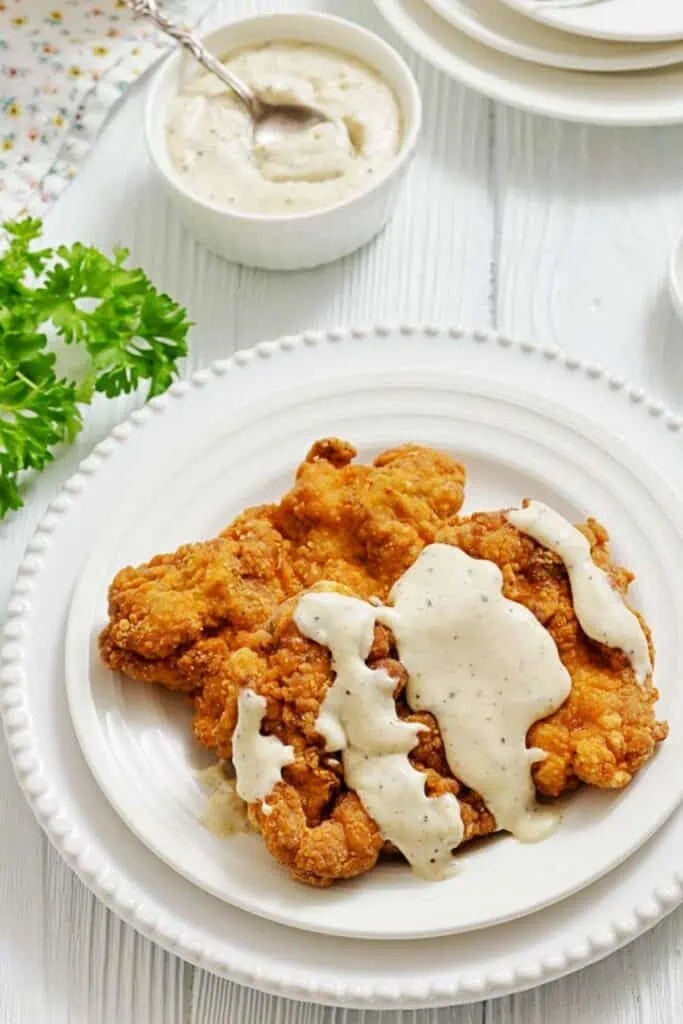 A plate of fried chicken topped with white gravy, served on a white plate with a bowl of gravy and parsley garnish nearby.