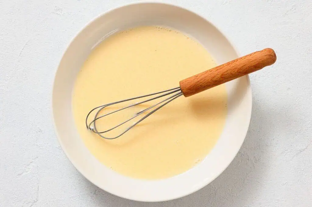 A metal whisk with a wooden handle rests in a white bowl containing a pale yellow liquid mixture on a light surface.