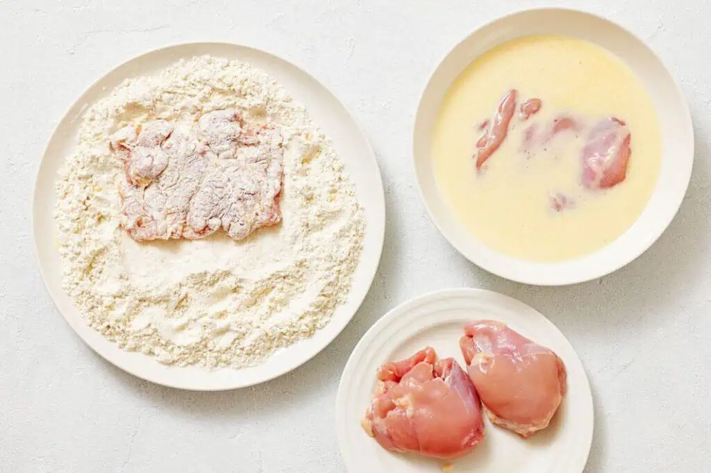 Three plates: one with raw chicken thighs, one with raw chicken in egg wash, and one with chicken pieces coated in flour.