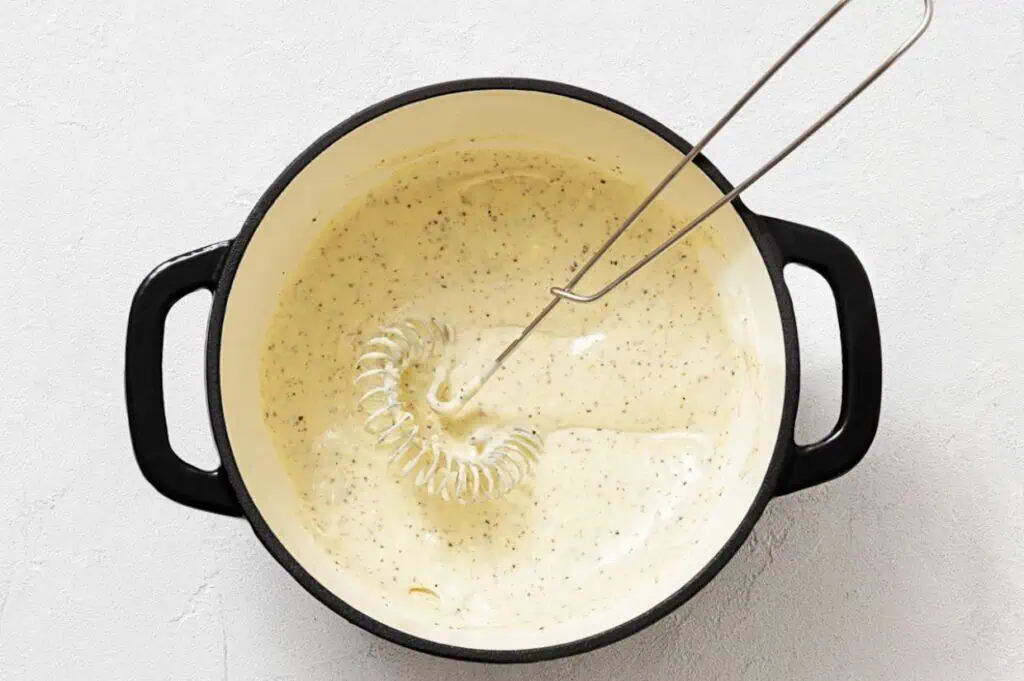 A metal whisk rests in a black pot filled with a creamy, seasoned sauce on a light-colored surface.