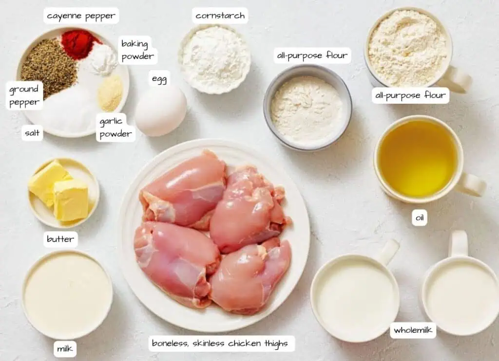 Assorted ingredients including boneless, skinless chicken thighs, spices, butter, flour, cornstarch, egg, oil, and milk arranged on a white surface, each labeled.