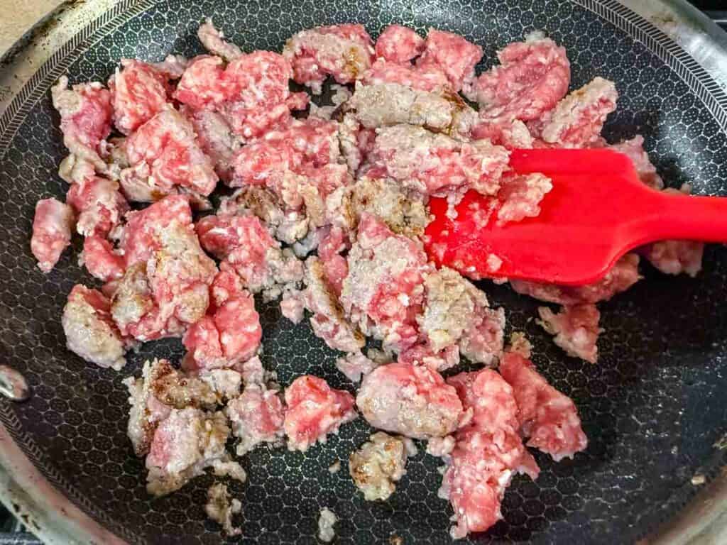 Ground beef being browned in a frying pan, with a red spatula stirring the meat.