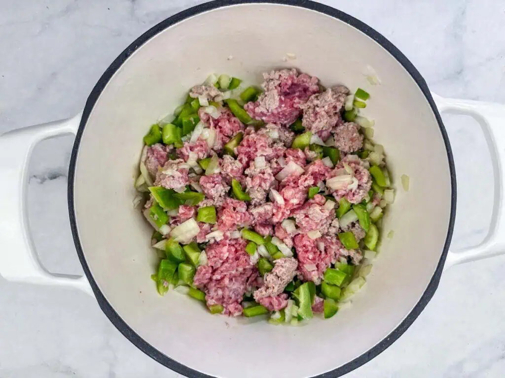 Raw ground meat, chopped green bell peppers, and diced onions in a white pot on a marble surface, ready to be cooked.