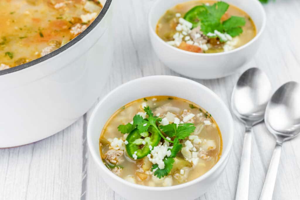 Three bowls of soup topped with cilantro, sliced jalape&ntilde;o, and cheese are on a light surface next to two spoons; a pot of soup is also partially visible.