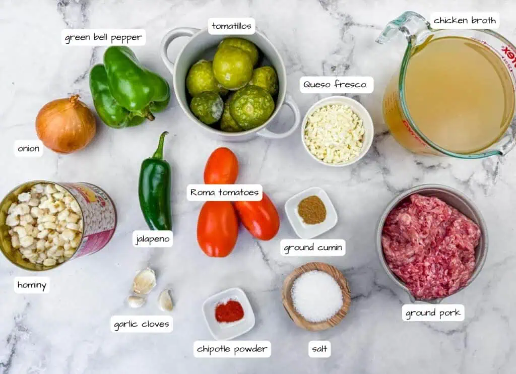 Ingredients for a recipe are arranged on a marble surface, including ground pork, hominy, green bell pepper, tomatillos, Roma tomatoes, onion, jalapeño, garlic, cheese, spices, and chicken broth.