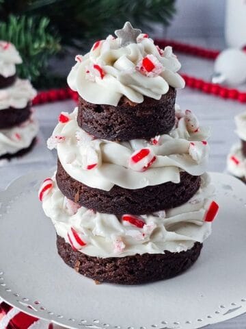 Three stacked brownie rounds with white frosting and crushed peppermint candies, arranged in the shape of Christmas trees on a white cake stand, with festive decorations in the background.
