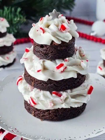 Three stacked brownie rounds with white frosting and crushed peppermint candies, arranged in the shape of Christmas trees on a white cake stand, with festive decorations in the background.