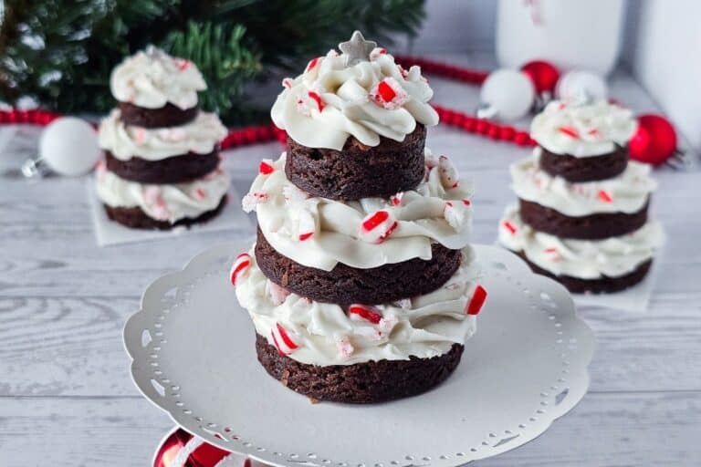 Three stacked brownie rounds with white frosting and crushed peppermint candies, arranged in the shape of Christmas trees on a white cake stand, with festive decorations in the background.