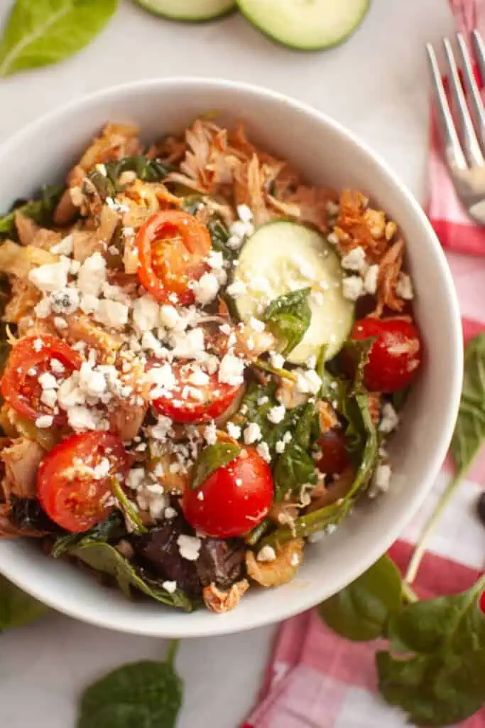 A white bowl filled with shredded chicken, cherry tomatoes, spinach, cucumber slices, and crumbled feta cheese, placed on a table with a fork and some spinach leaves nearby.