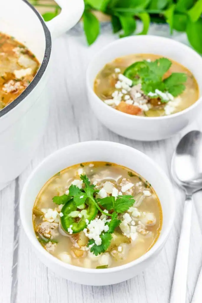 Bowls of white chili soup garnished with cilantro, jalapeño slices, and crumbled cheese sit on a light wooden surface next to a spoon and a white pot.