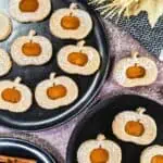 Assorted round cookies with pumpkin-shaped cutouts, dusted with powdered sugar, arranged on black plates with autumn-themed decorations nearby.