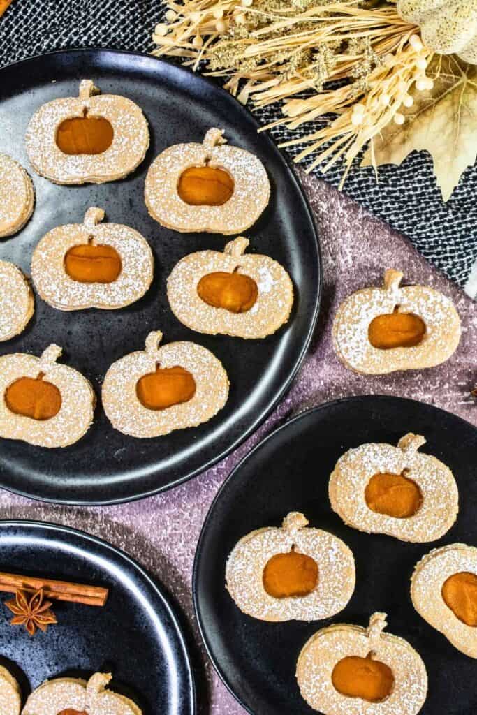 Assorted round cookies with pumpkin-shaped cutouts, dusted with powdered sugar, arranged on black plates with autumn-themed decorations nearby.