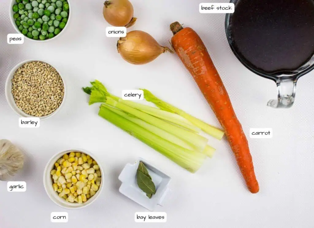 Assorted soup ingredients including peas, barley, corn, garlic, onions, celery, carrot, bay leaves, and a measuring cup of beef stock arranged on a white surface.