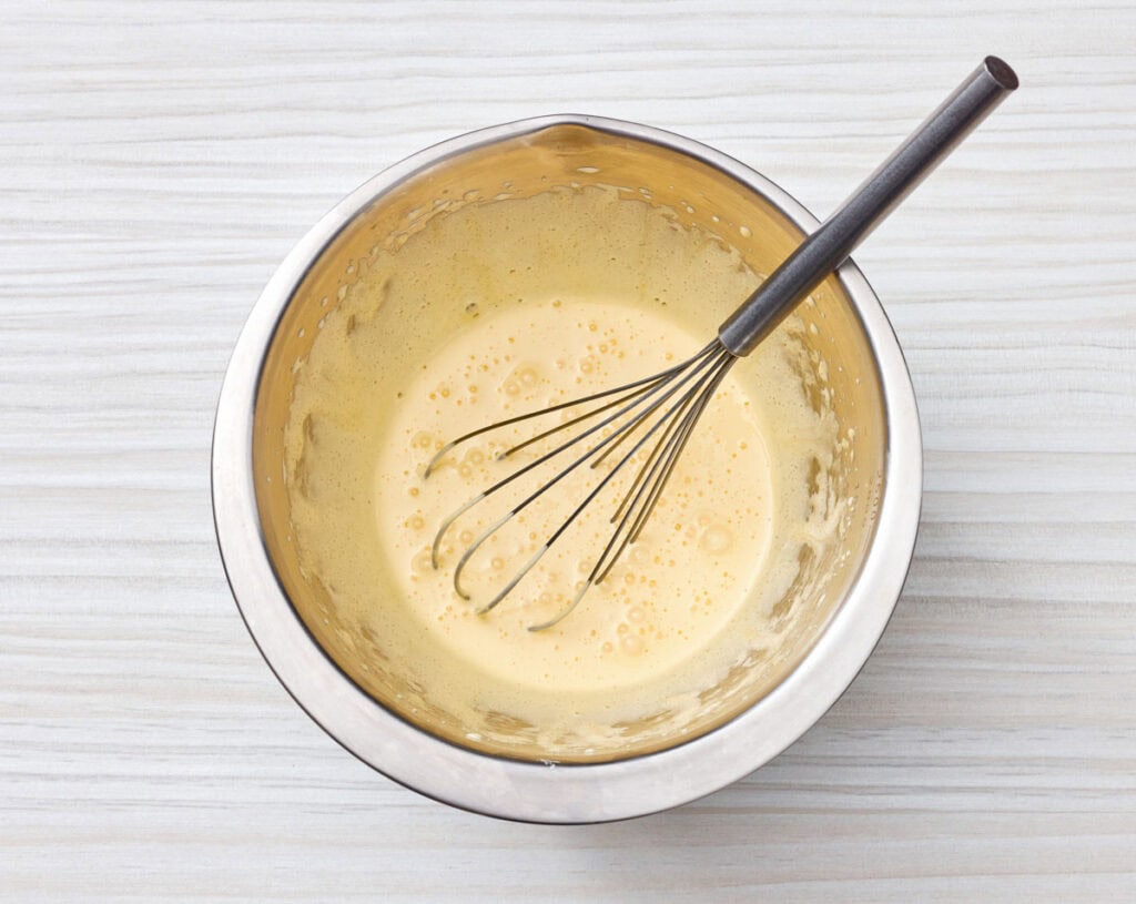 A metal mixing bowl with a whisk sits on a white surface, containing a pale, frothy batter or mixture.