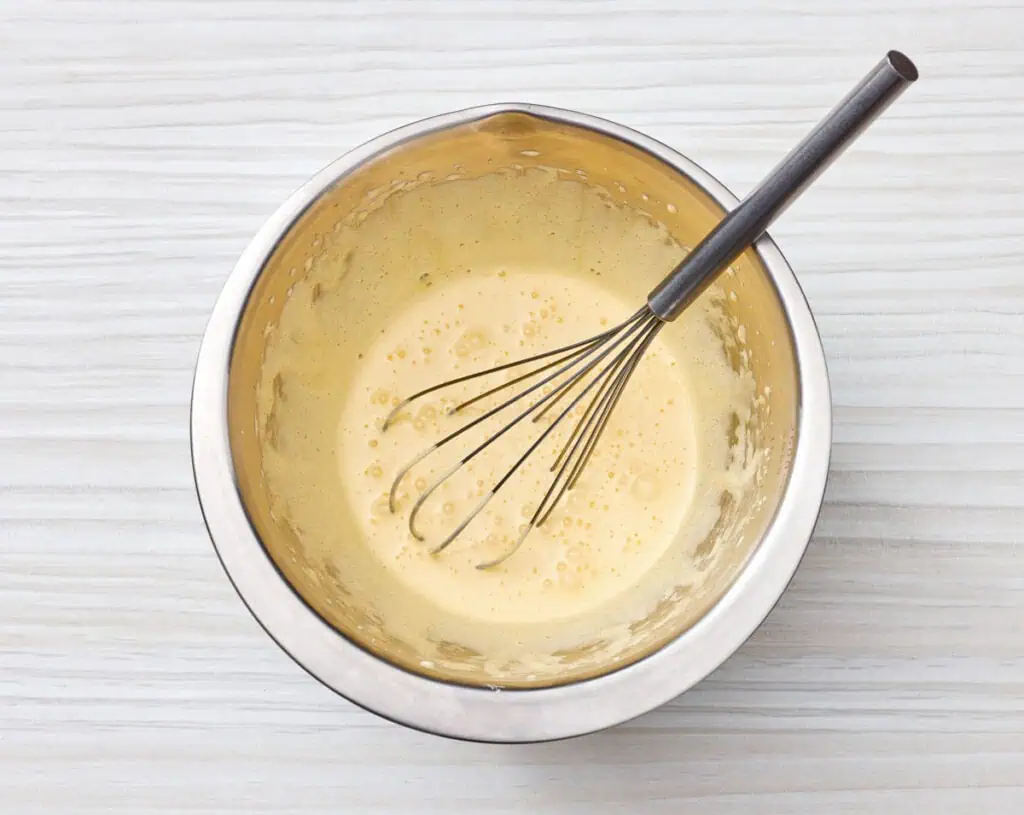A metal mixing bowl with a whisk sits on a white surface, containing a pale, frothy batter or mixture.