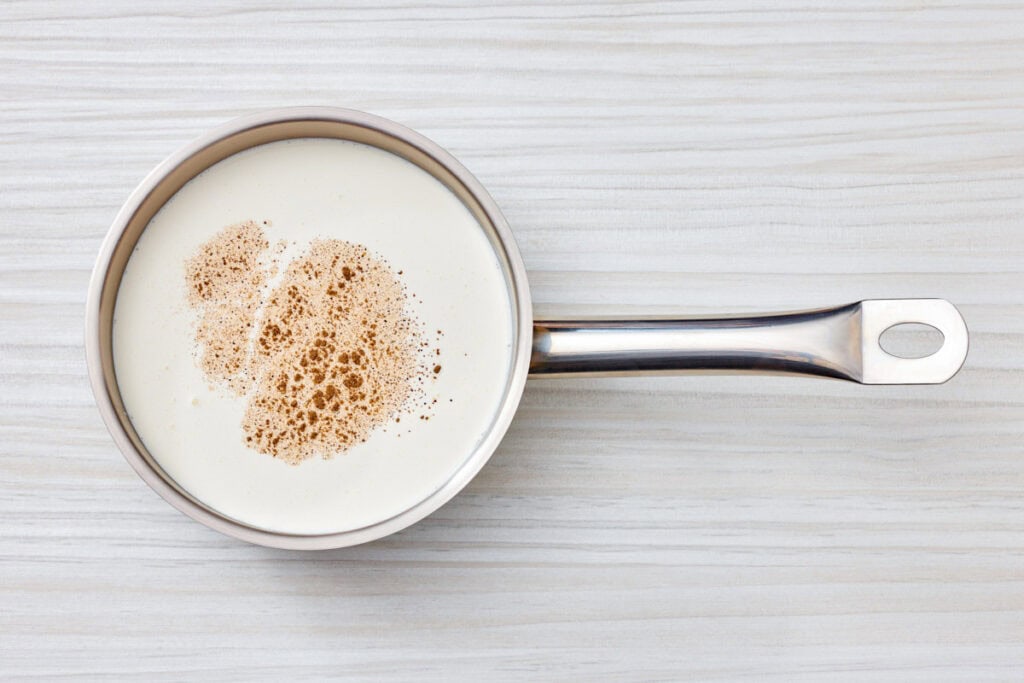 A stainless steel saucepan filled with cream and a sprinkle of ground nutmeg on top, placed on a light wood surface.