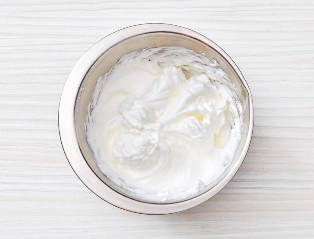 A metal mixing bowl filled with stiffly beaten egg whites on a light-colored wooden surface.