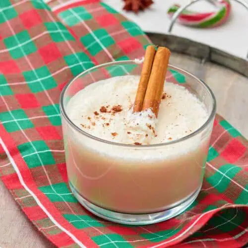 A glass of eggnog topped with ground nutmeg and two cinnamon sticks, placed on a red and green plaid napkin on a wooden tray.