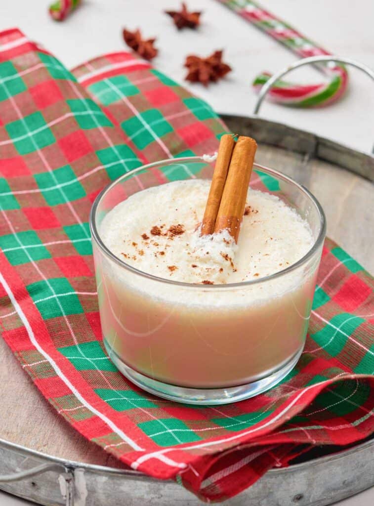 A glass of eggnog topped with ground nutmeg and two cinnamon sticks, placed on a red and green plaid napkin on a wooden tray.